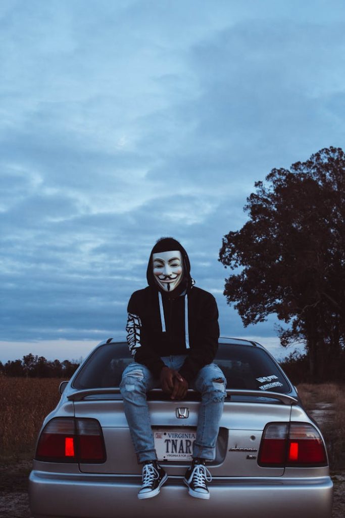 Person in a hacker mask sits on a car outdoors in Williamsburg, VA.